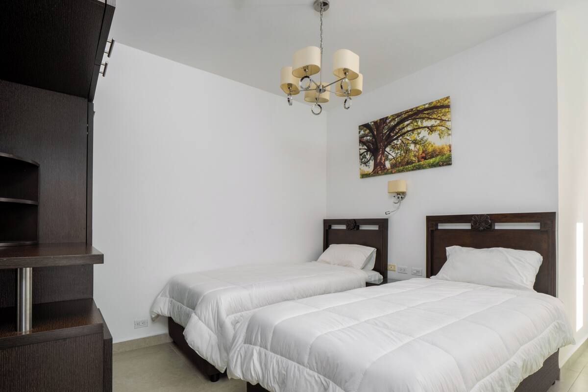 A hotel room with two beds, a dark wooden dresser, a wall-mounted lamp, and a painting of a tree.