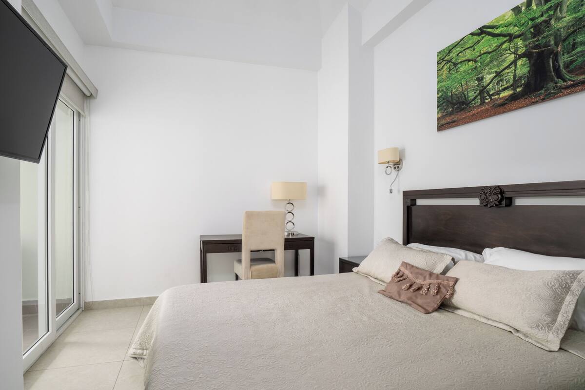 A hotel room with a large bed, a desk with a chair, a lamp, and a painting of a forest on the wall.