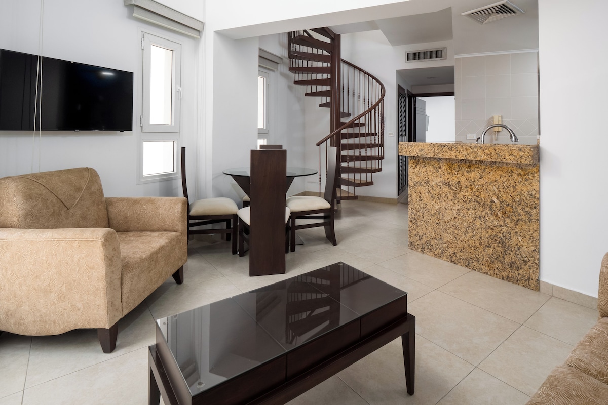 A modern living room with a sofa, a coffee table, a TV, a dining table with chairs, and a kitchen area with a granite countertop and a spiral staircase.