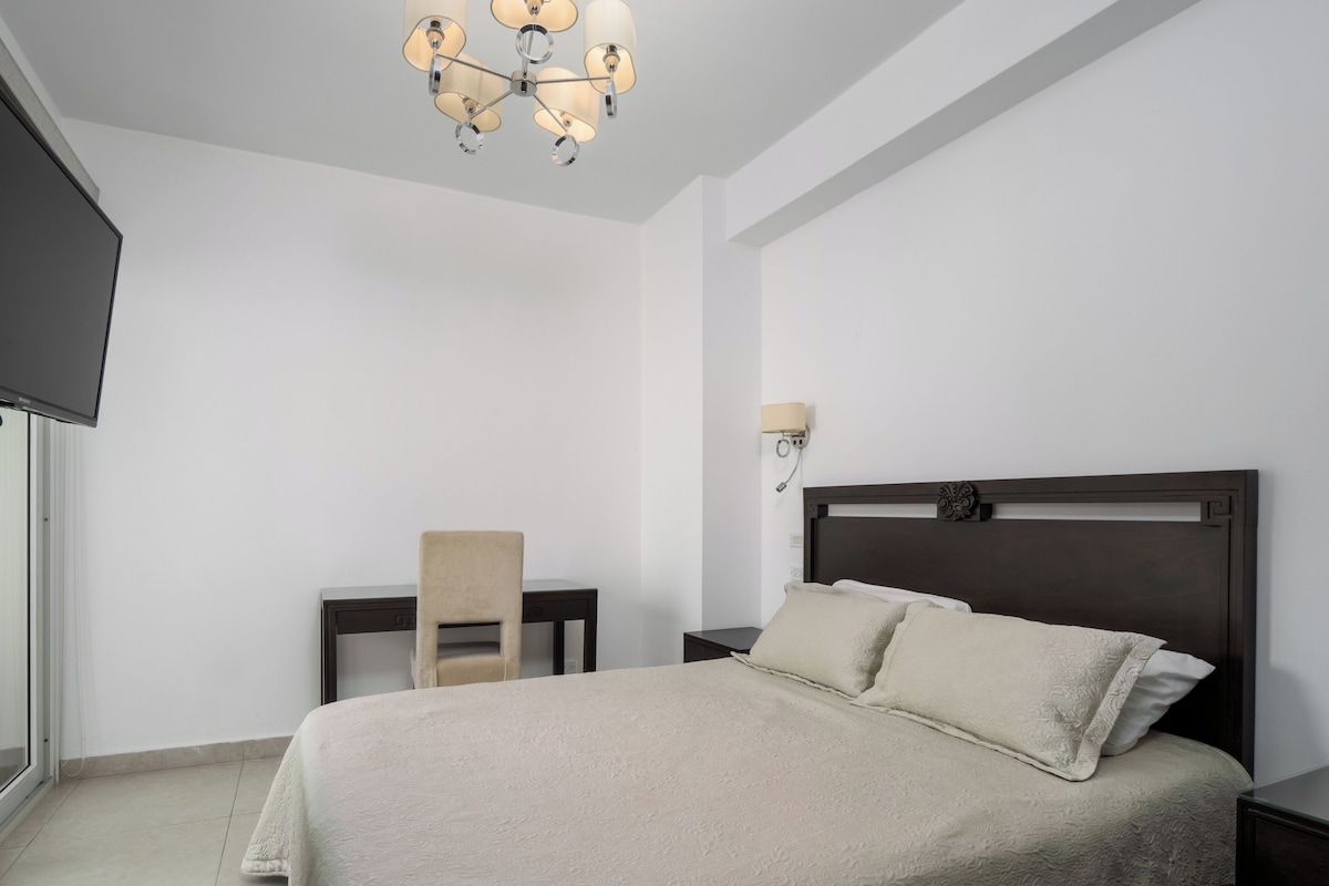 A bedroom with a bed, a chair, a desk, a wall-mounted lamp, and a television.