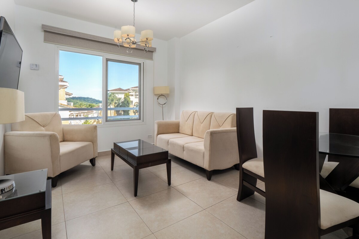 A modern living room with a sofa, armchairs, a coffee table, and a TV. A large window offers a view of buildings and greenery.