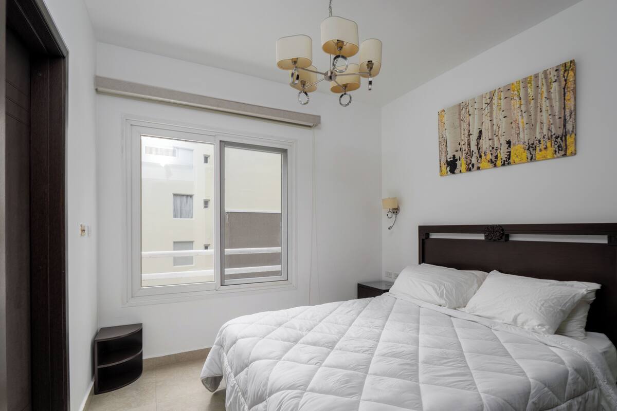A bedroom with a bed, a window, a wall-mounted lamp, and a painting.
