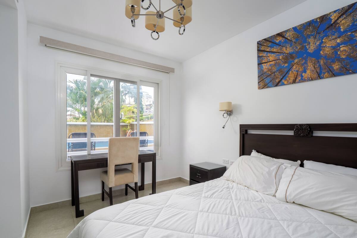 A hotel room with a large bed, a desk with a chair, a nightstand, a wall-mounted lamp, and a window with a view of the outdoors.