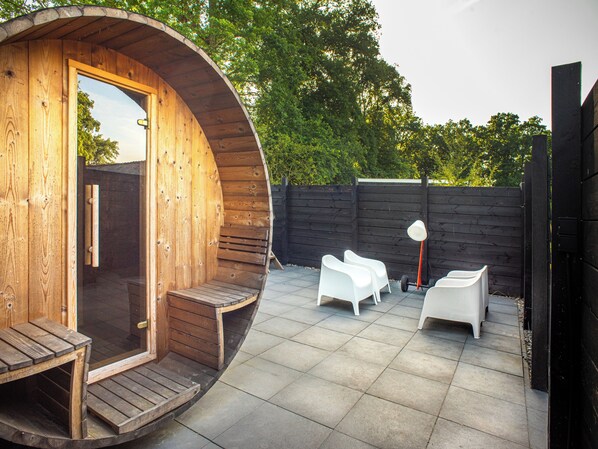 A wooden structure with a curved entrance, a small outdoor seating area with white chairs, and a paved patio.