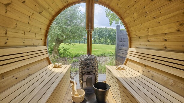 Una sauna con interior de madera, calentador de piedra y vistas a un paisaje verde a través de un amplio ventanal circular.