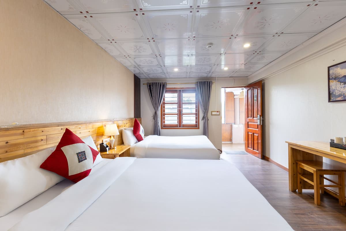 A hotel room with two beds, wooden headboards, a wooden table, and a view of the outside through a window with curtains.