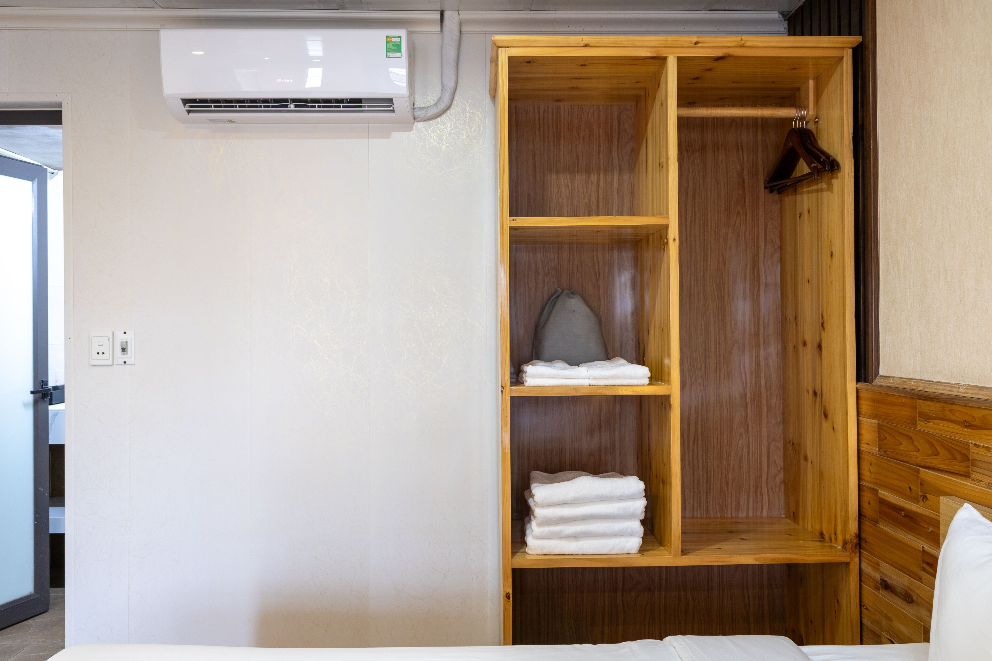 A hotel room with a bed, a wooden wardrobe with shelves, and an air conditioning unit.
