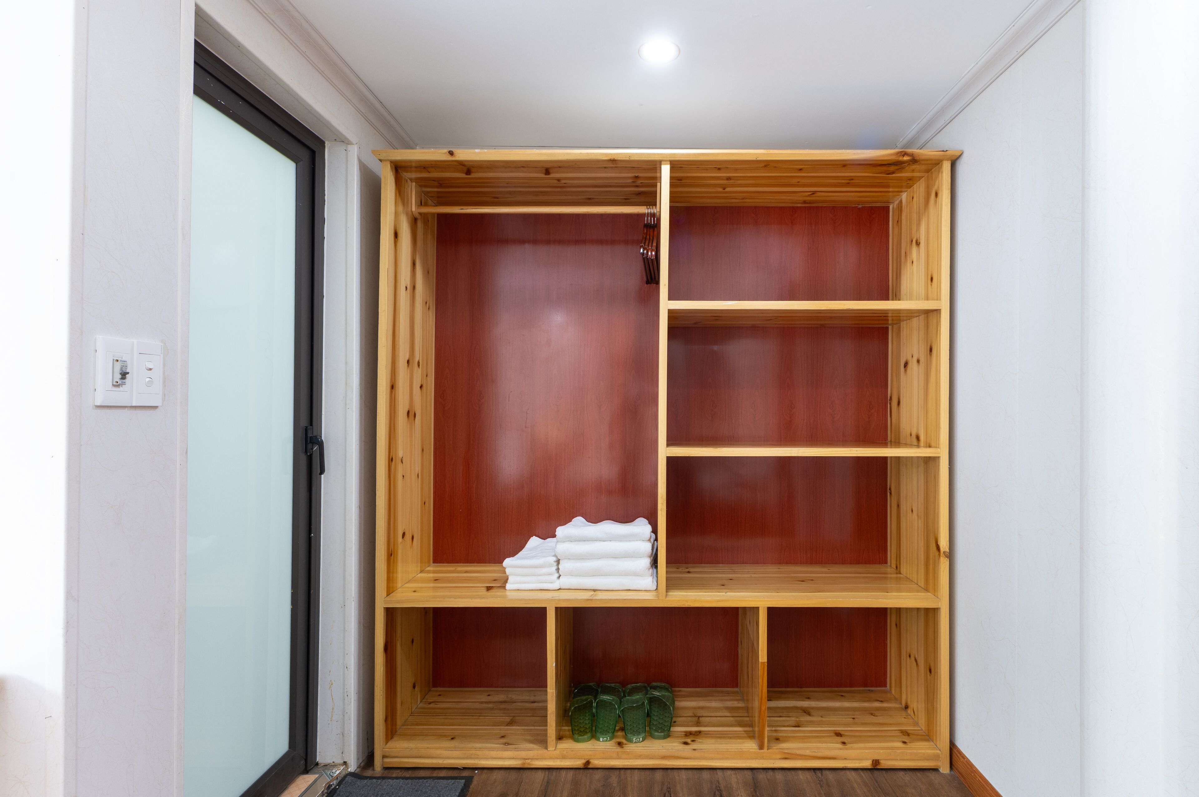 Guarda-roupa de madeira com várias prateleiras e uma porta, num quarto com parede de cor clara e uma tomada elétrica visível.