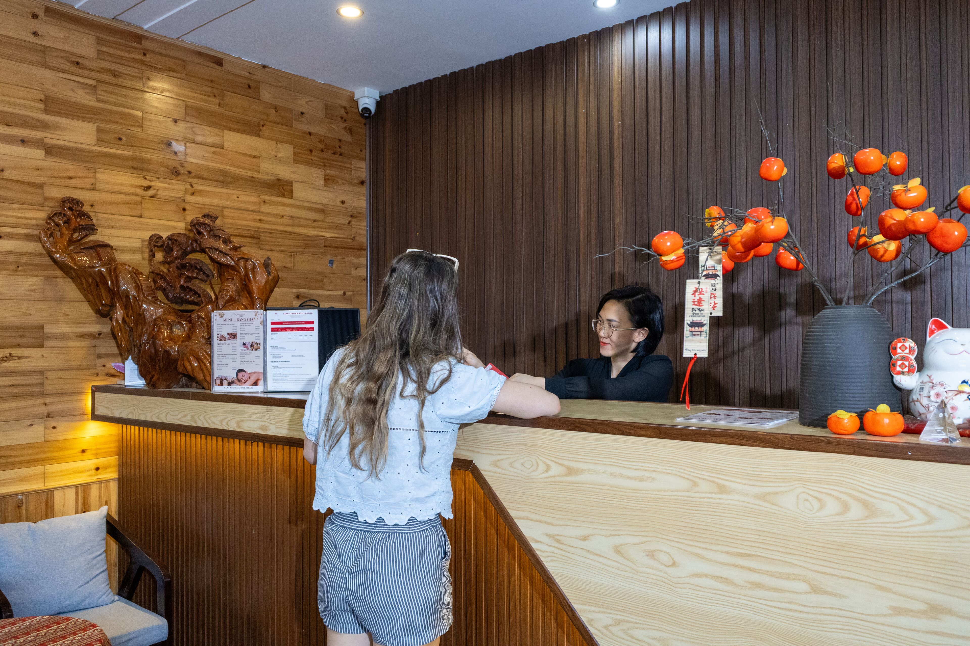 Un comptoir d’accueil avec un mur revêtu de panneaux en bois, une réceptionniste et un vase décoratif contenant des oranges.