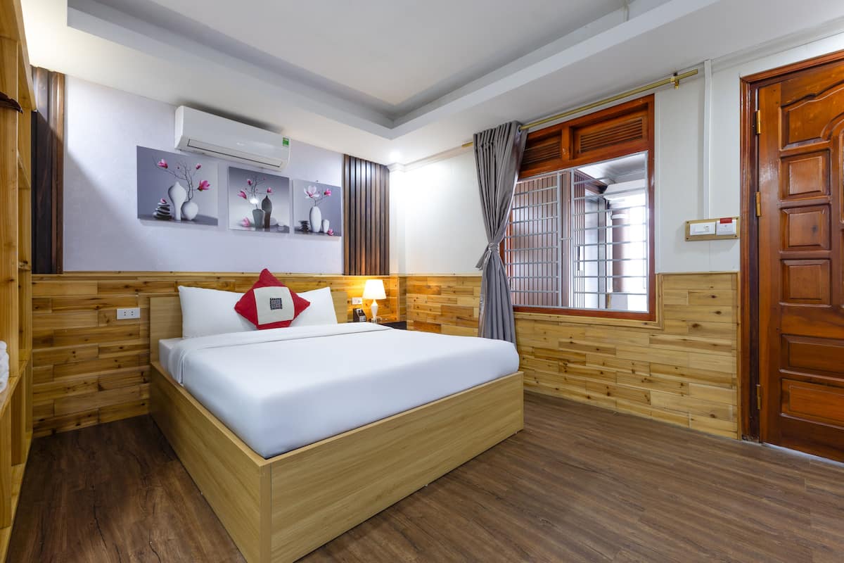 A hotel room with a wooden bed, a nightstand, a wall-mounted air conditioner, and a window with a view of a building.