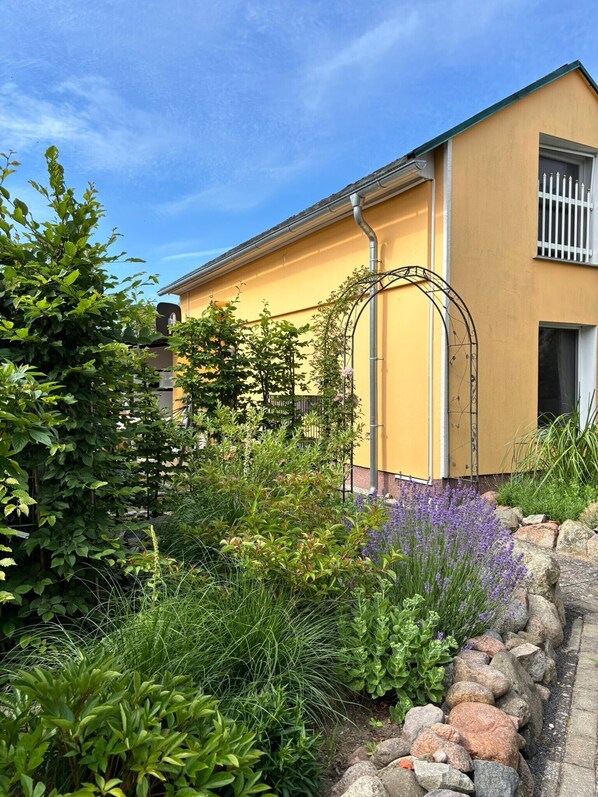 A yellow building with a metal archway, surrounded by a garden with various plants and a stone border.