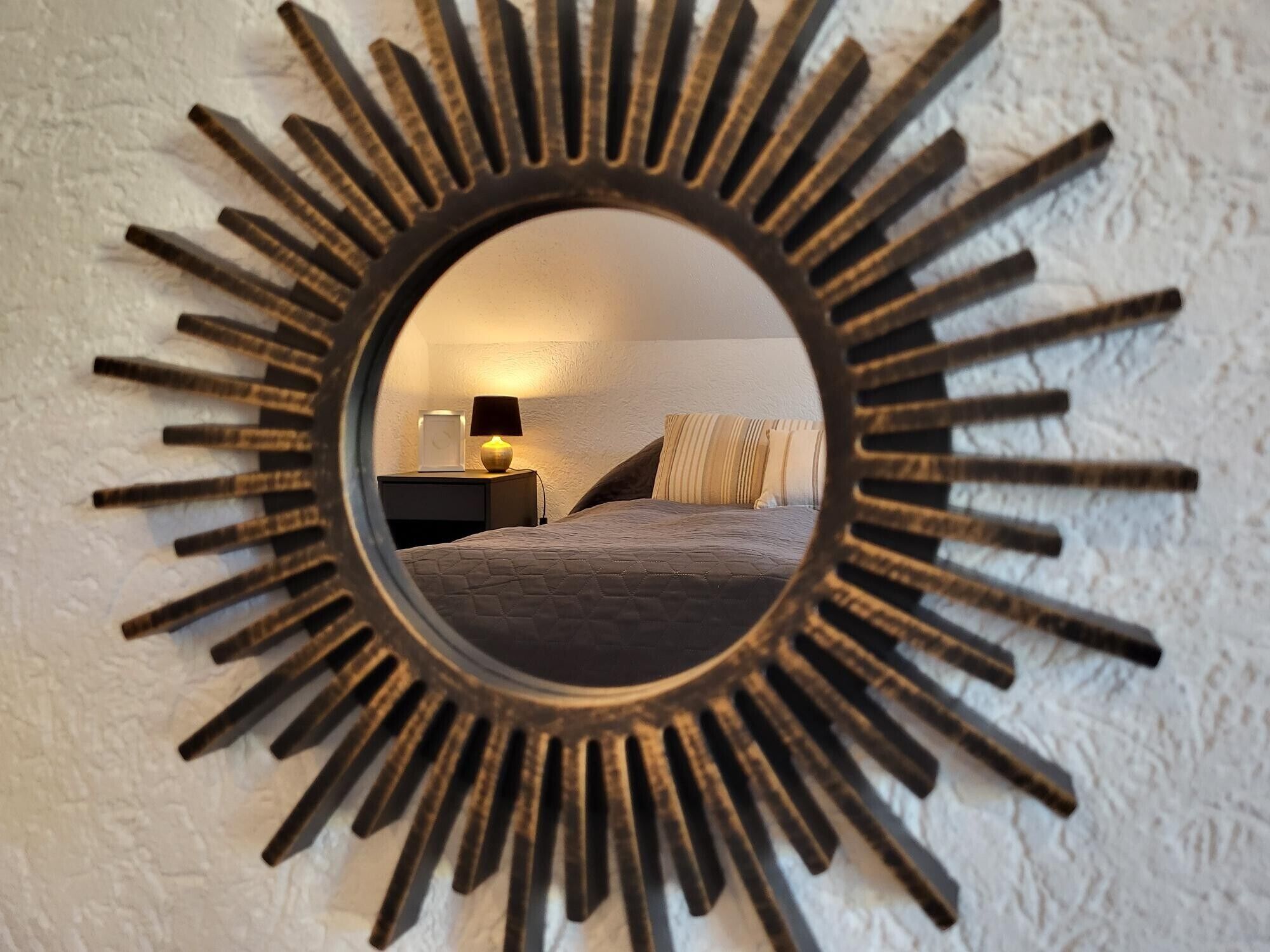A sunburst mirror reflecting a bedroom with a bed, pillows, a nightstand, and a lamp.