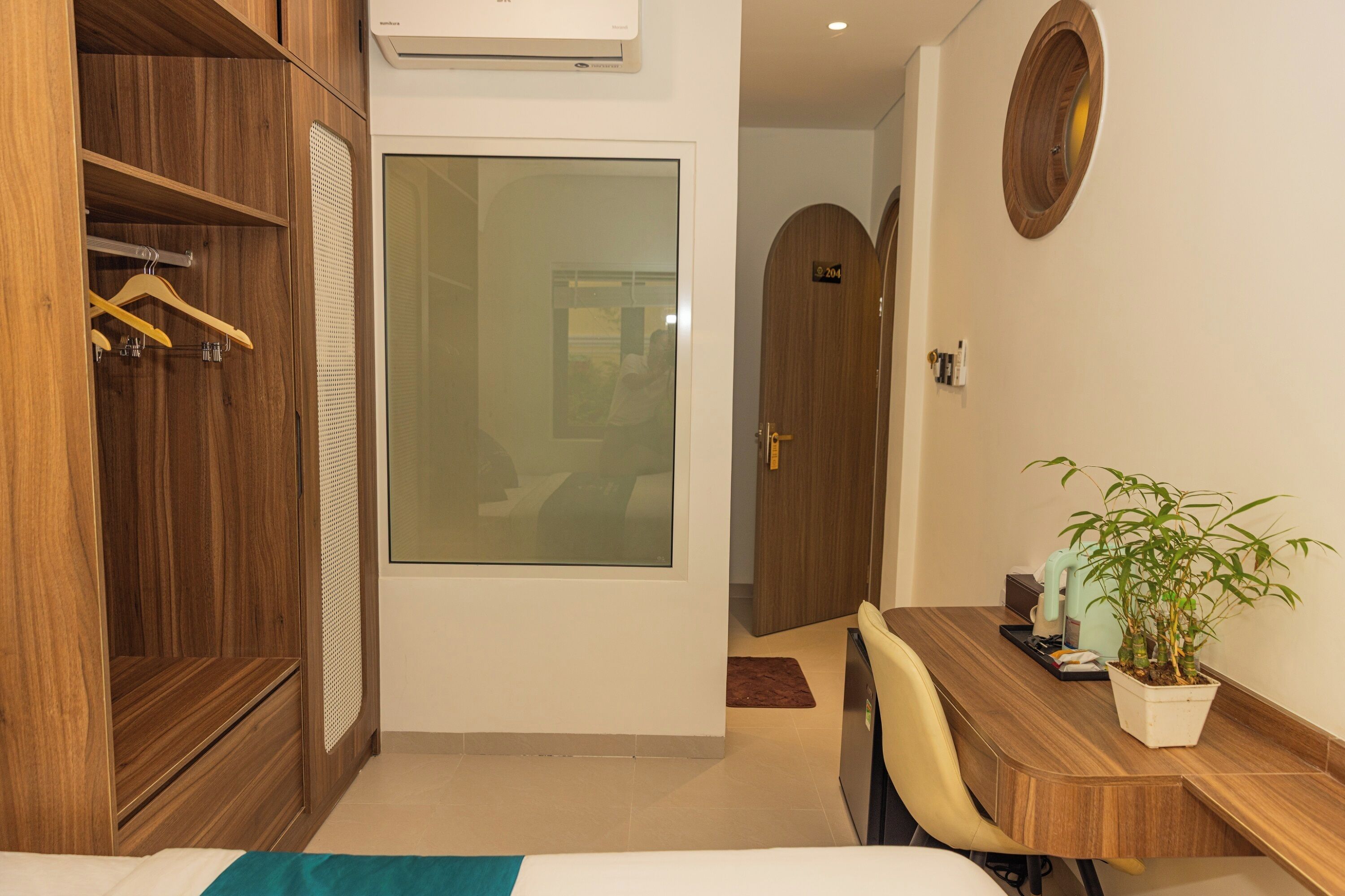 A hotel room with a wooden wardrobe, a bed, a desk with a chair, a potted plant, and a door with a number sign.
