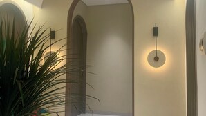 Um corredor com piso branco, paredes bege e teto com iluminação embutida. Há duas grandes portas arqueadas e um vaso de planta à esquerda.