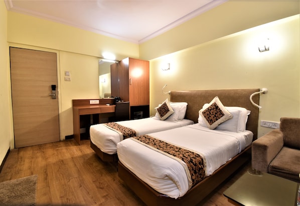 A hotel room with two beds, a wooden wardrobe, a desk with a mirror, and a brown armchair. - Hotel Atlantic (Mumbai)