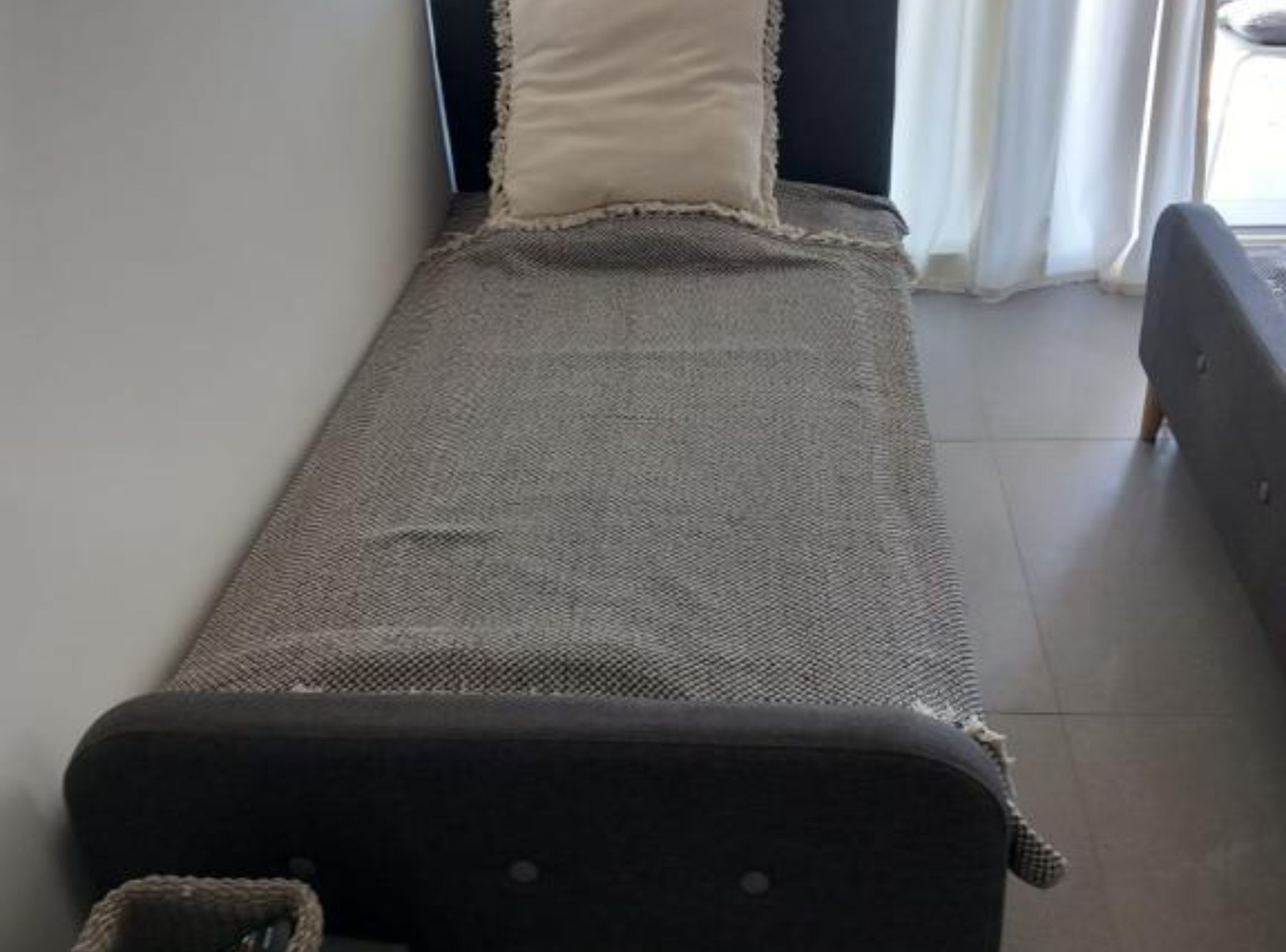 A single bed with a grey textured bedspread and a white pillow, positioned against a wall with a window and curtains.