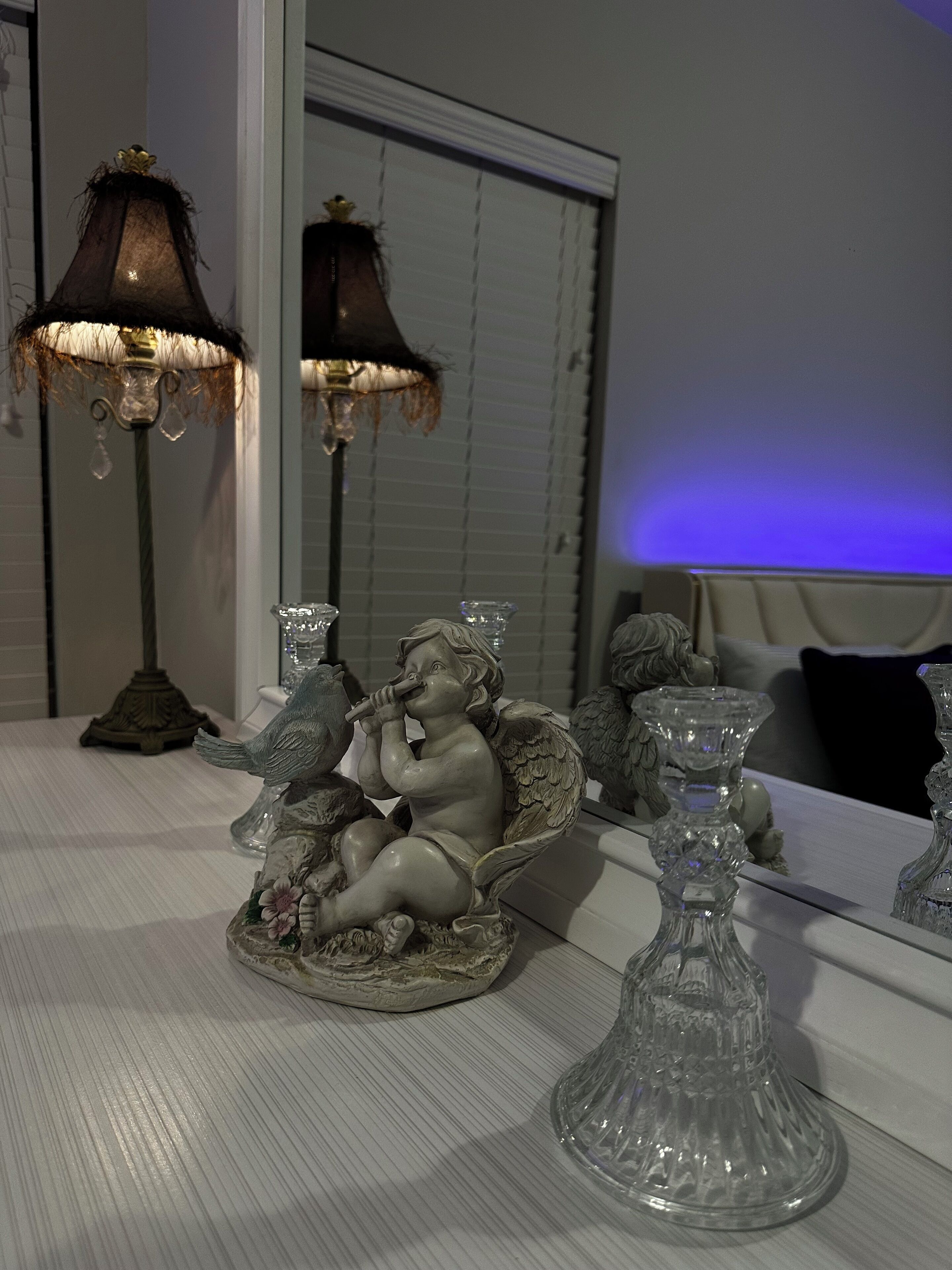 A table with a decorative angel figurine, a lamp, and a mirror reflecting a window with blinds.