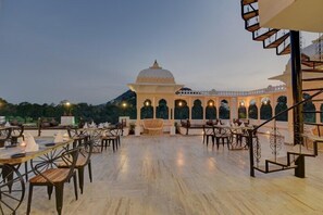 An outdoor dining area with tables and chairs, a central seating area with a wooden bench, and a view of a traditional building with arches and a dome. - janak vilas (Udaipur)