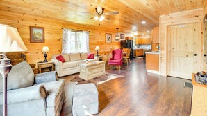 A wooden interior with a living area featuring a sofa, armchairs, a coffee table, and a dining table. A ceiling fan and a kitchen with a refrigerator are also visible. - 18 Mi to Red River Gorge: Beattyville Retreat (Beattyville)