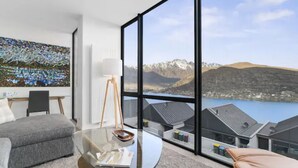 Ein modernes Wohnzimmer mit einem Glastisch, einem grauen Sofa und einem großen Fenster mit Blick auf Berge und einen See.