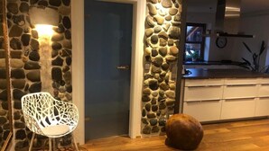 A room with a stone wall, a white chair, a round wooden object, and a kitchen area in the background.