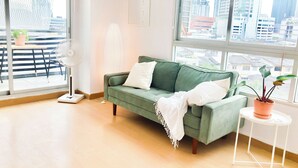Una sala de estar moderna con un sofá verde, cojines blancos, un ventilador de pie, una mesita con una planta en maceta y vistas a la ciudad a través de amplios ventanales.