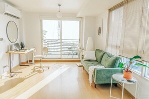 Un salon moderne avec un canapé vert, une petite table ronde ornée d’une plante, un bureau avec un ordinateur portable et une grande fenêtre équipée de stores.
