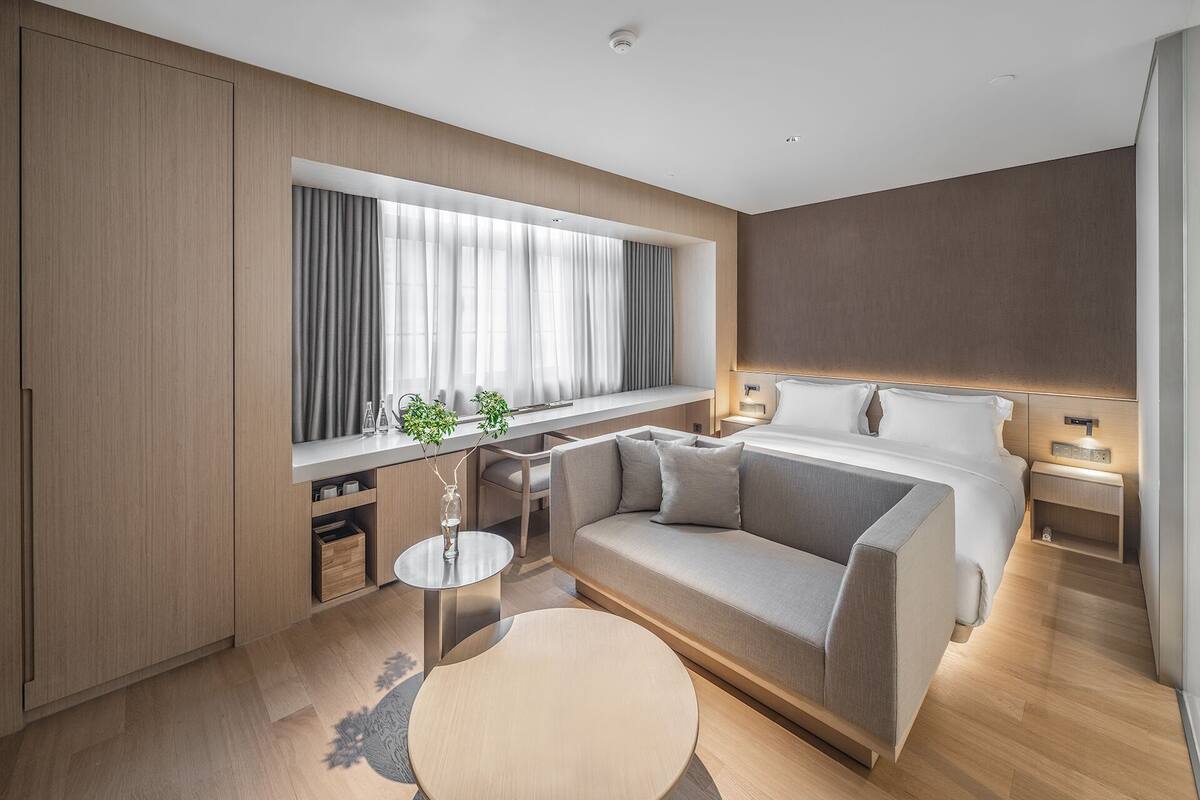 Une chambre d’hôtel moderne avec un canapé, une table basse ronde, un lit avec une tête de lit et une fenêtre avec des rideaux.