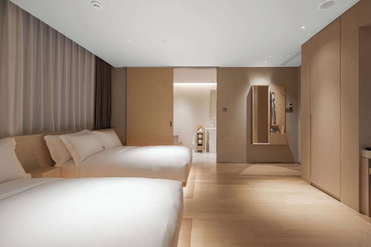 Une chambre d’hôtel moderne avec un grand lit, un plus petit lit, une armoire en bois et une salle de bain visible à travers une porte ouverte.