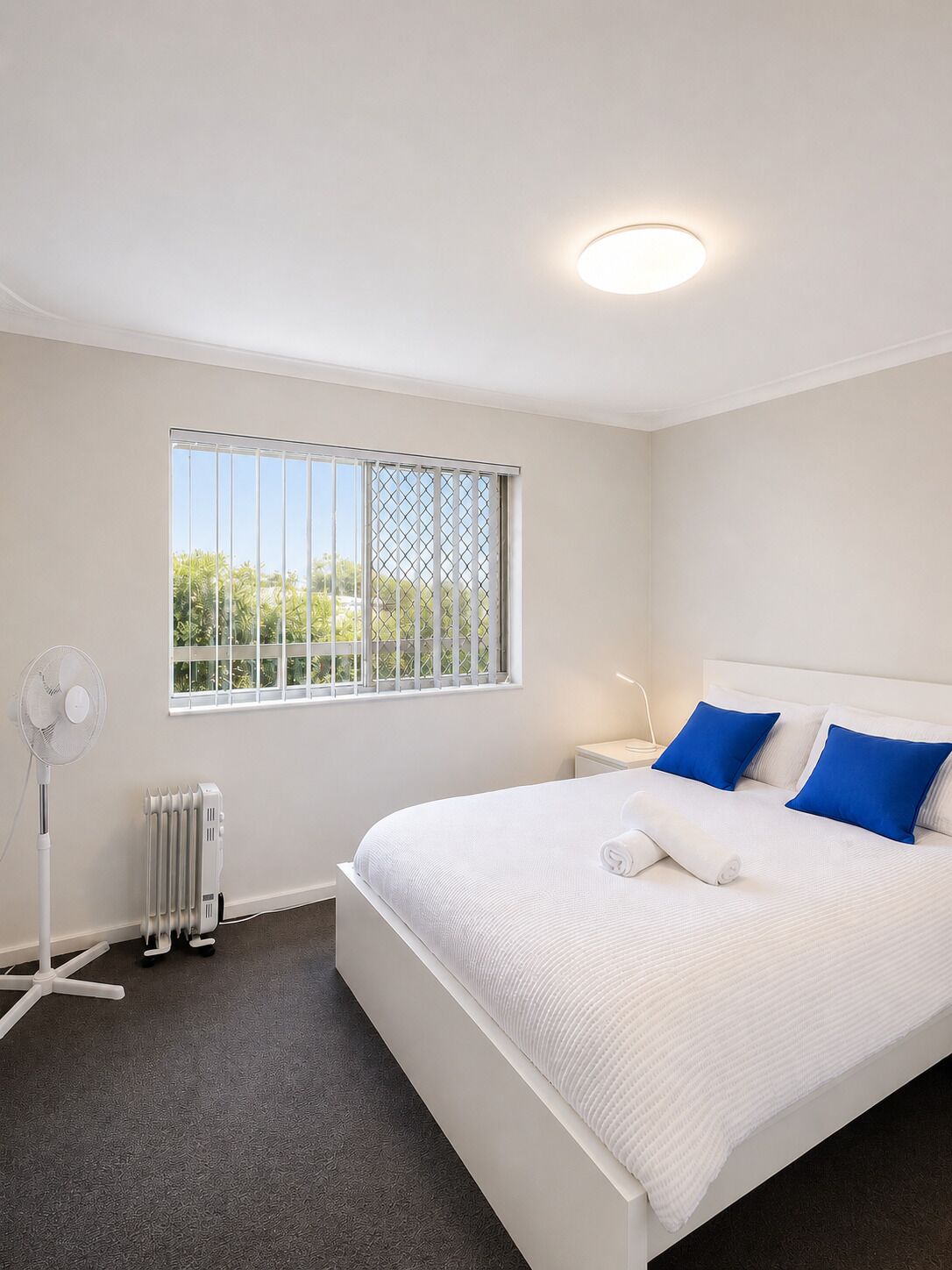 Une chambre à coucher avec un lit, une fenêtre avec des stores, un ventilateur sur pied et un radiateur.