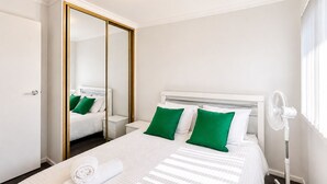 Ein Hotelzimmer mit einem großen Bett, einem Nachttisch, einem Spiegel, einem Standventilator und einem Fenster.