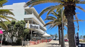 Ein Gebäude direkt am Strand mit einem Schild für das Restaurant 'The Sandbar', Palmen und einem wolkenlosen, blauen Himmel.