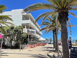 Un bùtiment situé en bord de mer, avec une enseigne indiquant le restaurant « The Sandbar », des palmiers et un ciel bleu azur.