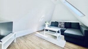 Un espacio habitable moderno estilo loft, con techo inclinado, televisor de pantalla plana sobre un soporte blanco, sofá gris oscuro con cojines estampados y una mesa de centro blanca.