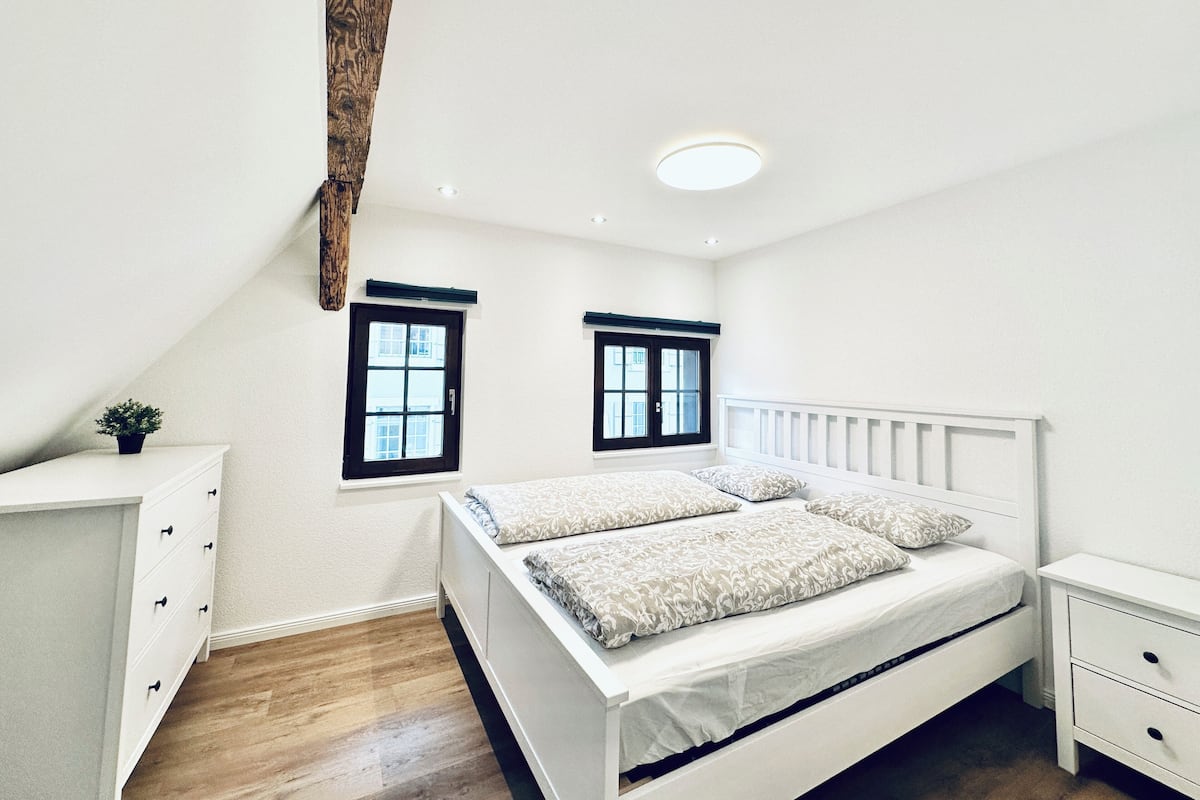 A bedroom with a white bed, two windows, a white dresser, and a wooden beam on the ceiling.