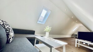 A modern loft bedroom with a sloped ceiling, a skylight, a grey sofa, a white table, and a TV stand. - Business Apartment Wangen (Wangen an der Aare)