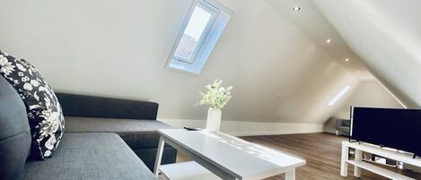 A modern loft bedroom with a sloped ceiling, a skylight, a grey sofa, a white table, and a TV stand.