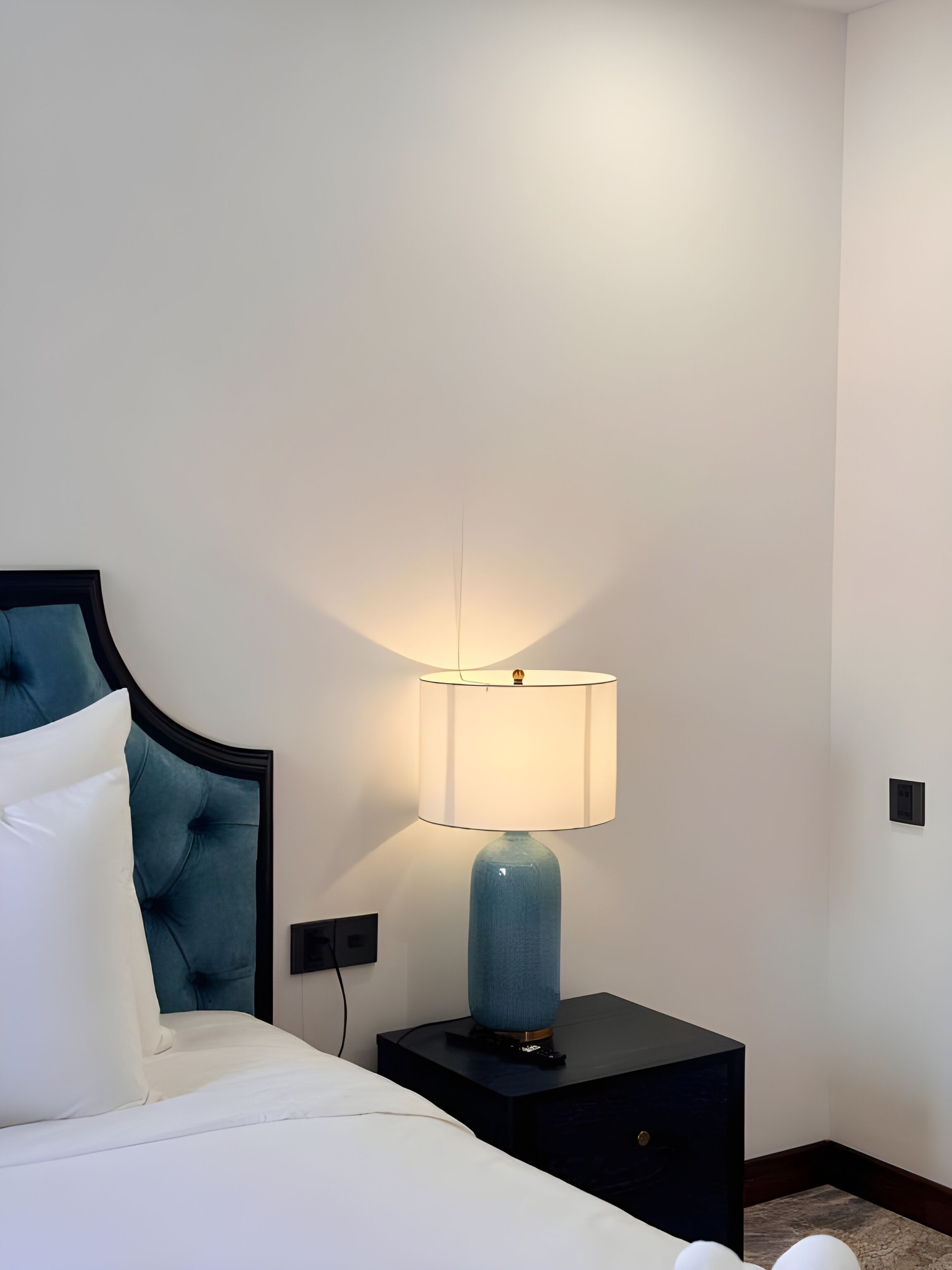 Un letto rifatto con lenzuola bianche e una testiera blu, una lampada blu su un comodino nero e una parete bianca con una presa elettrica.