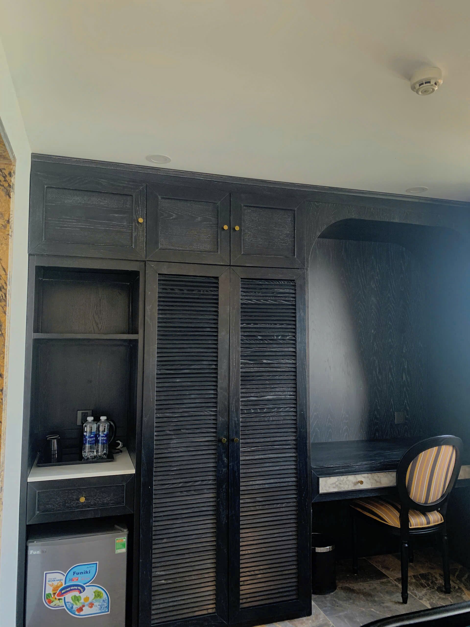 Een kamer met een zwarte garderobe, een bureau en een stoel. Er staan twee flessen op het onderste plankje van de garderobe.