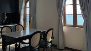 A dining area with a marble table and chairs, a view of the sea through large windows, and a kitchen in the background.