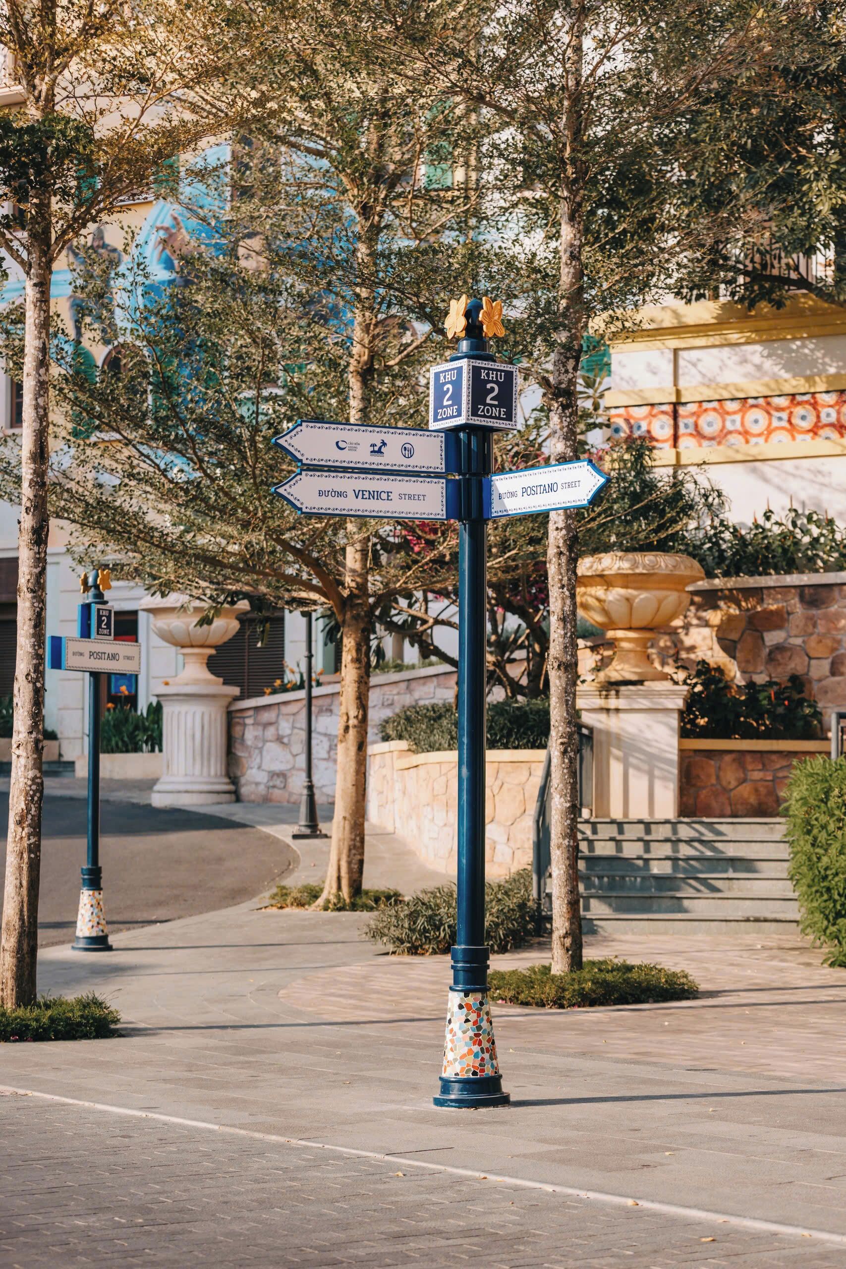 A street sign with directions to Duong Venice Street and Postano Street, surrounded by trees and a building with a blue and orange facade.