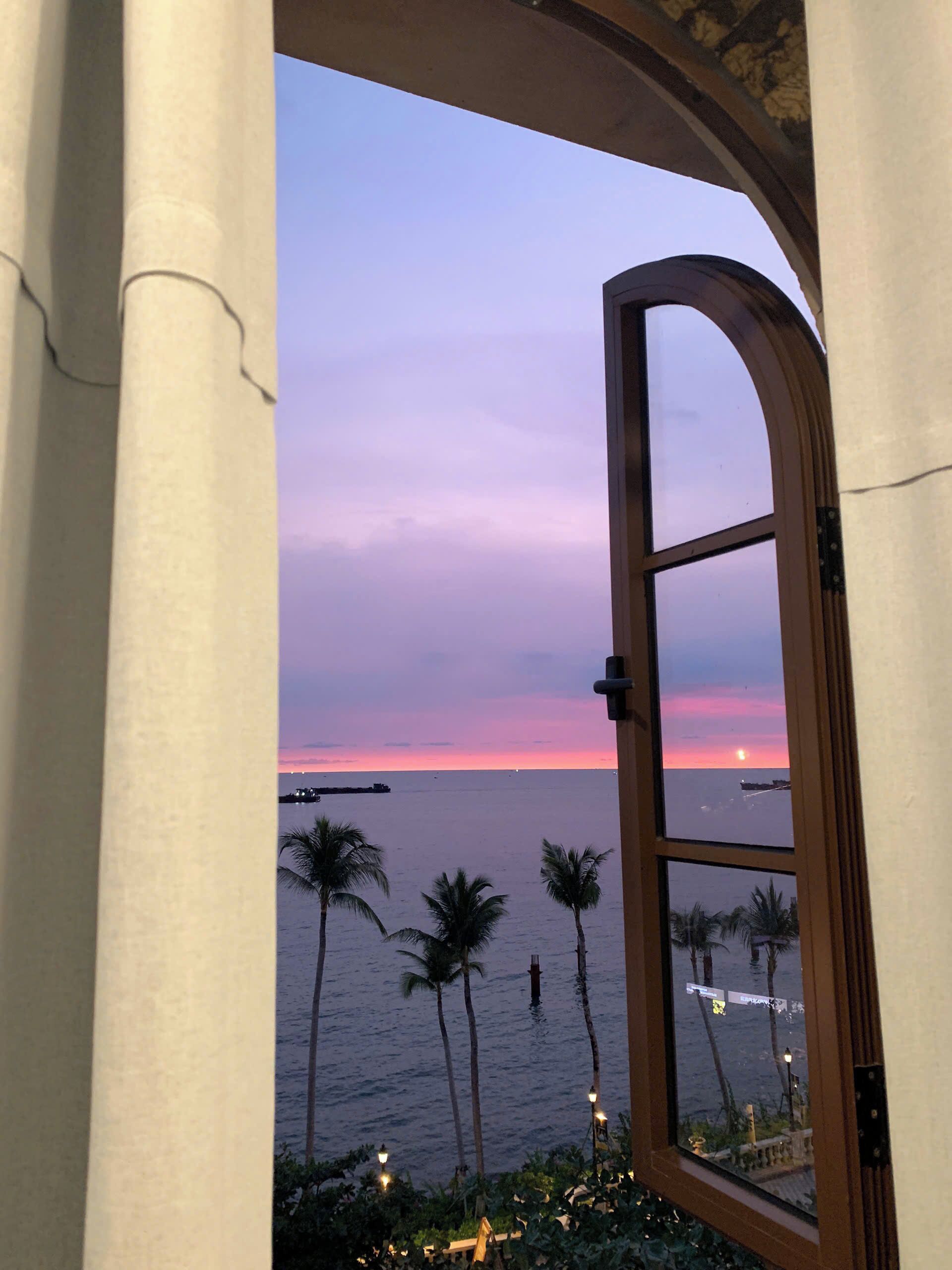 Vista da janela com moldura de madeira, com um corpo d'água, palmeiras e um pôr do sol.