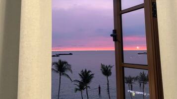 Vista da janela com moldura de madeira, com um corpo d'água, palmeiras e um pôr do sol.
