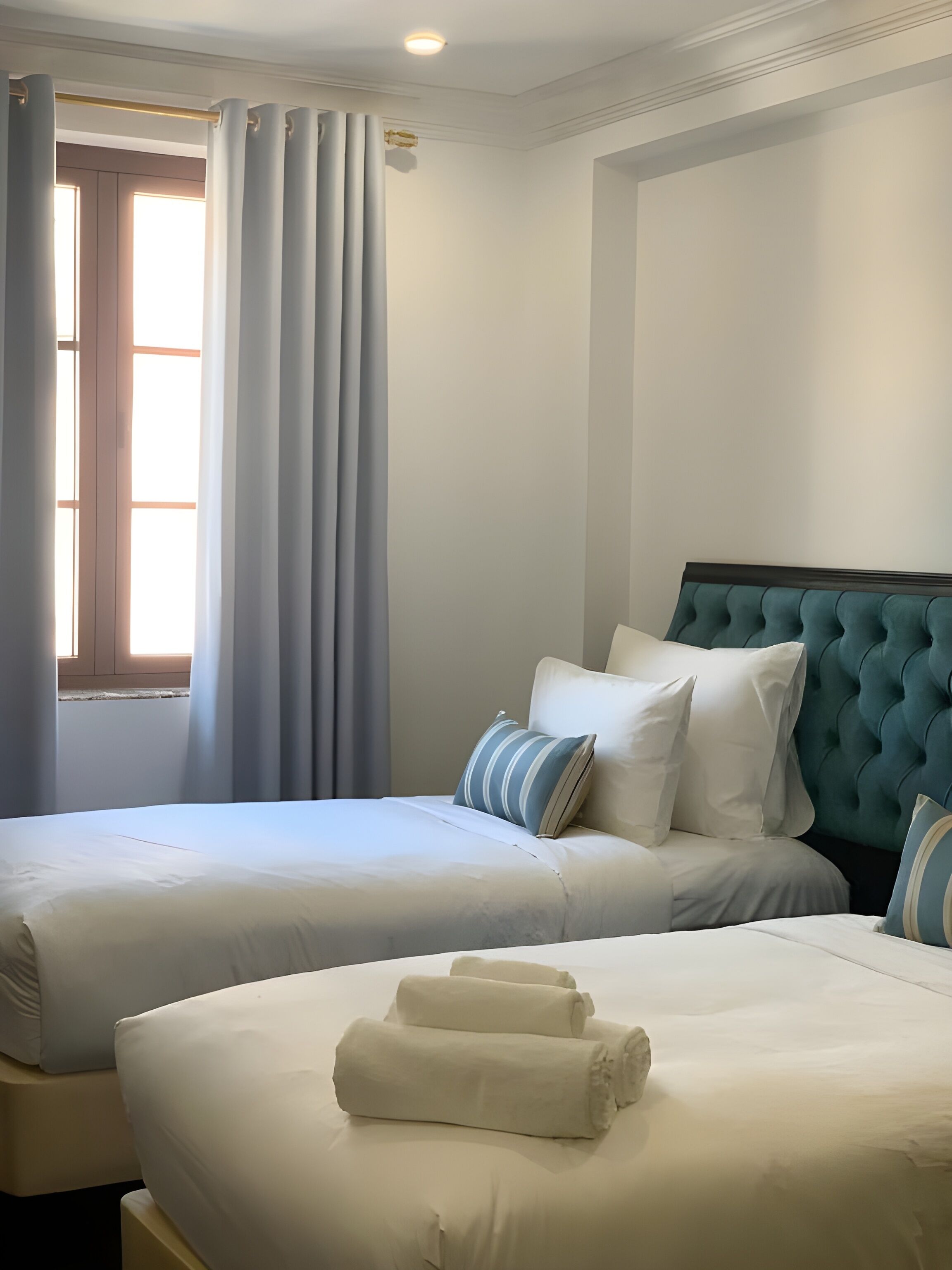 Una habitación de hotel con dos camas, un ventanal con cortinas y un cabecero capitonado.