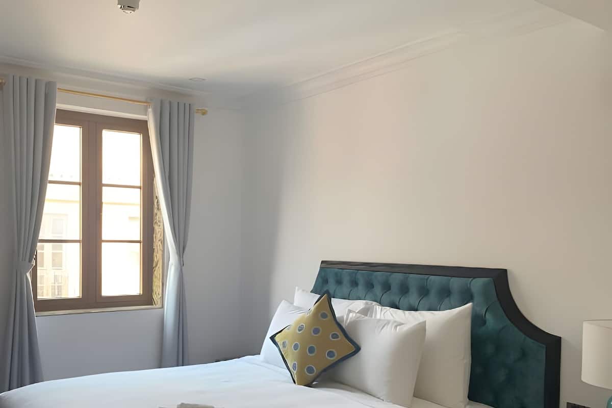 Un lit bien fait, avec du linge de lit blanc et une serviette pliée, une tête de lit rembourrée bleue et une fenêtre ornée de rideaux bleu clair.