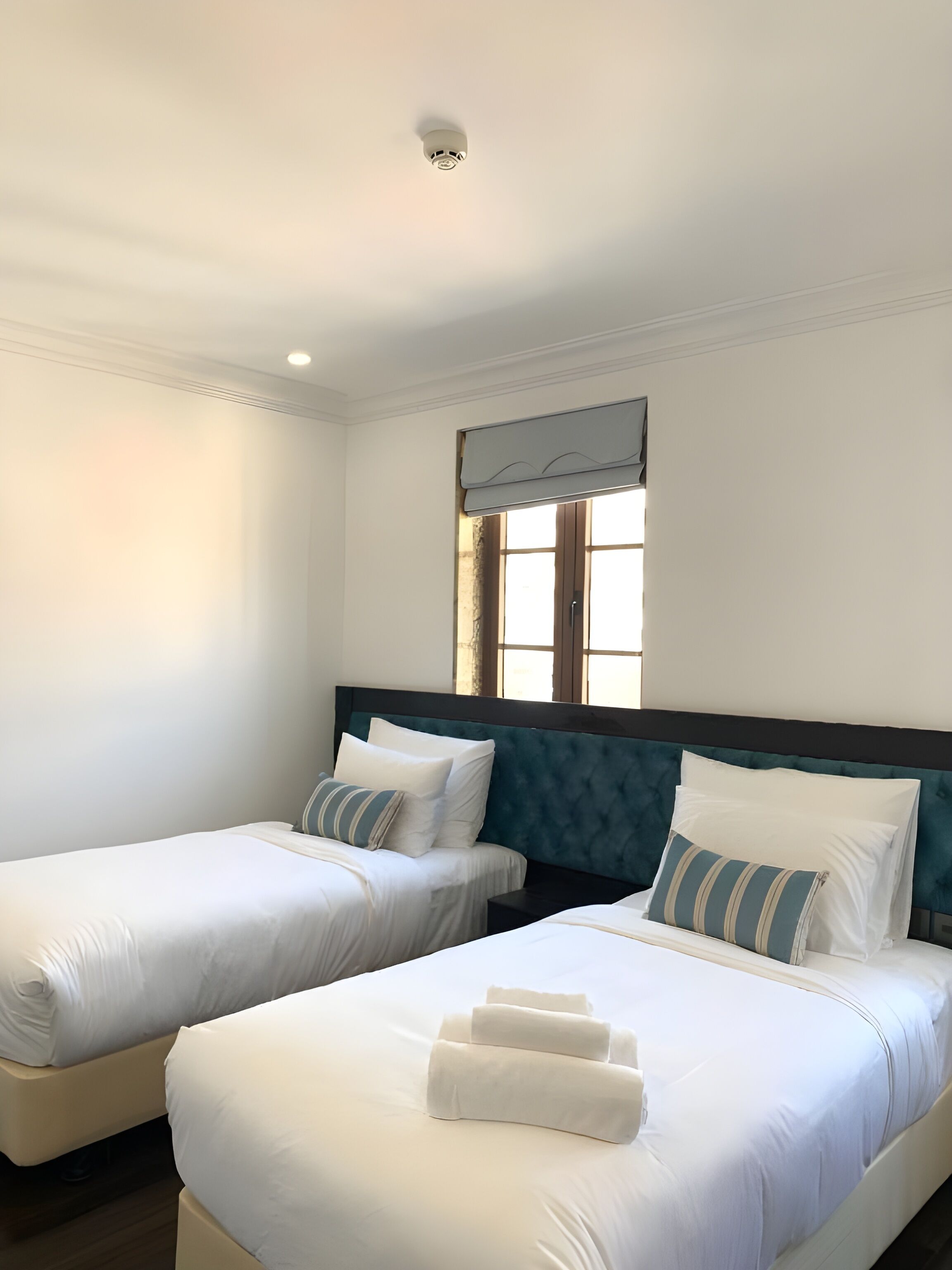 Habitación de hotel con dos camas, una ventana con persianas, una lámpara de techo y un cabecero oscuro.