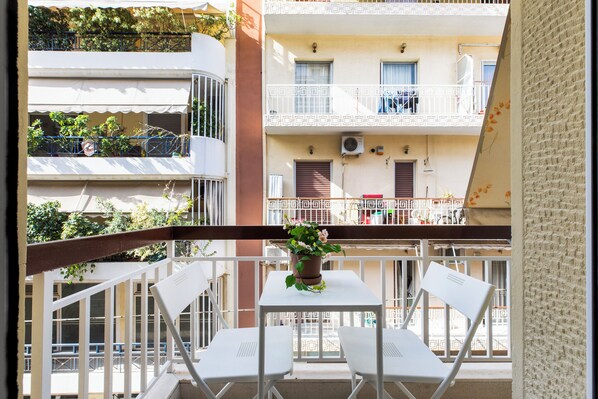 A balcony with white chairs and a table, a potted plant, and a view of a multi-story building with balconies and windows.