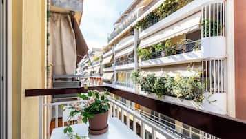 A balcony with a potted plant, white railing, and a view of multi-story buildings with balconies and plants.