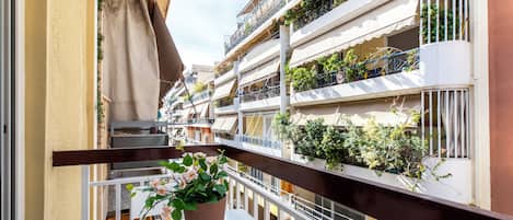 A balcony with a potted plant, white railing, and a view of multi-story buildings with balconies and plants.