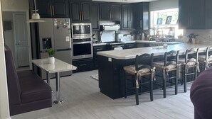 A modern kitchen with dark cabinets, stainless steel appliances, a central island with bar stools, and a dining area with a white table and bench. - Charming 4-br Wine Country Retreat — Walk Downtown Solvang + BBQ & Fireplace (Solvang)